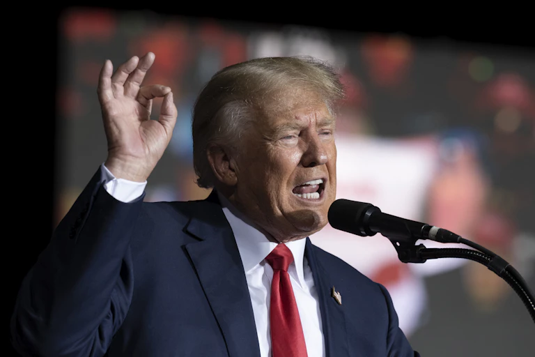 A white man in a blue suit holds his right hand up pincing his thumb and index finger speaks in front of a microphone