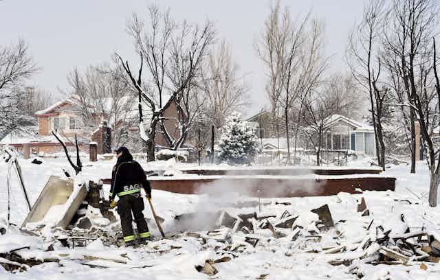 A firefighter stands with an ax at a hotspot of a home that burned to the foundation. Three houses appear untouched across the street.