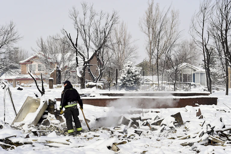 A firefighter stands with an ax at a hotspot of a home that burned to the foundation. Three houses appear untouched across the street.