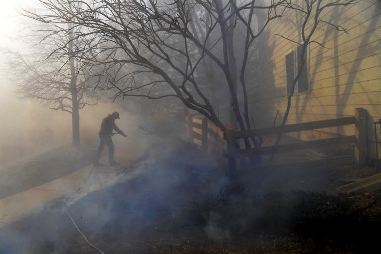 A man uses a garden hose to try to put out flames in the yard of a home. Smoke rises from the ground about 10 feet from the house.