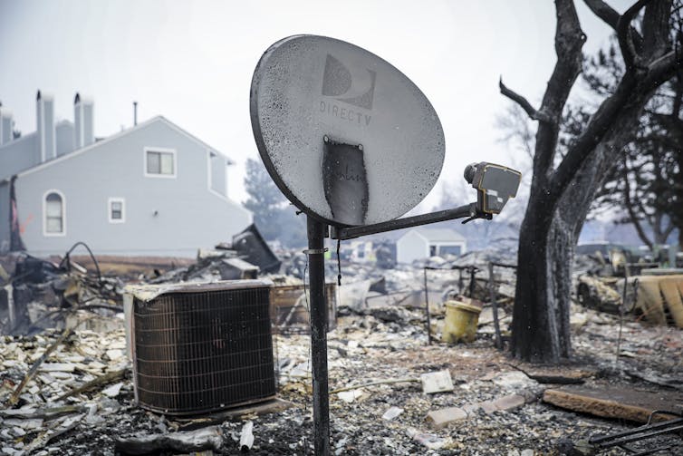 All that remains of a house is its satellite dish and air conditioner on an ashy lot. Other homes are still standing in the background.
