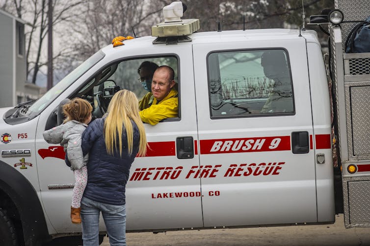 A woman holds a young girl on her hip as she talks with firefighters who are sitting in a truck with