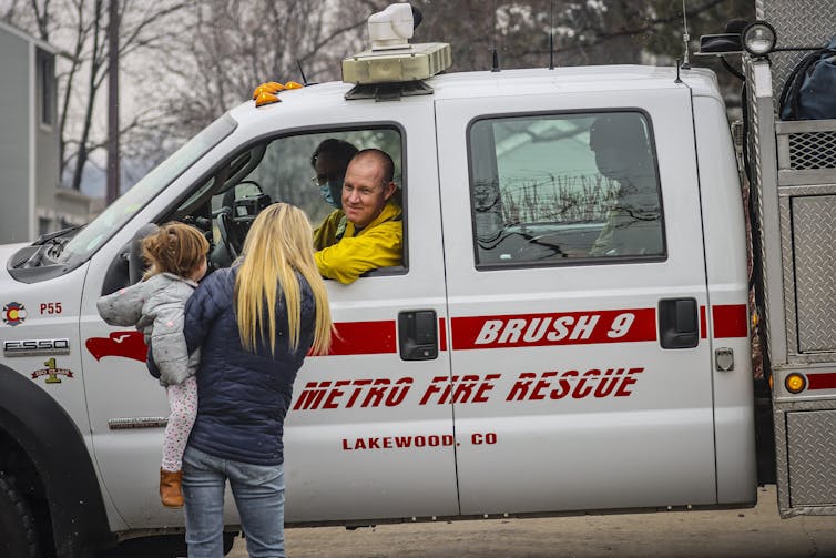 A woman holds a young girl on her hip as she talks with firefighters who are sitting in a truck with 