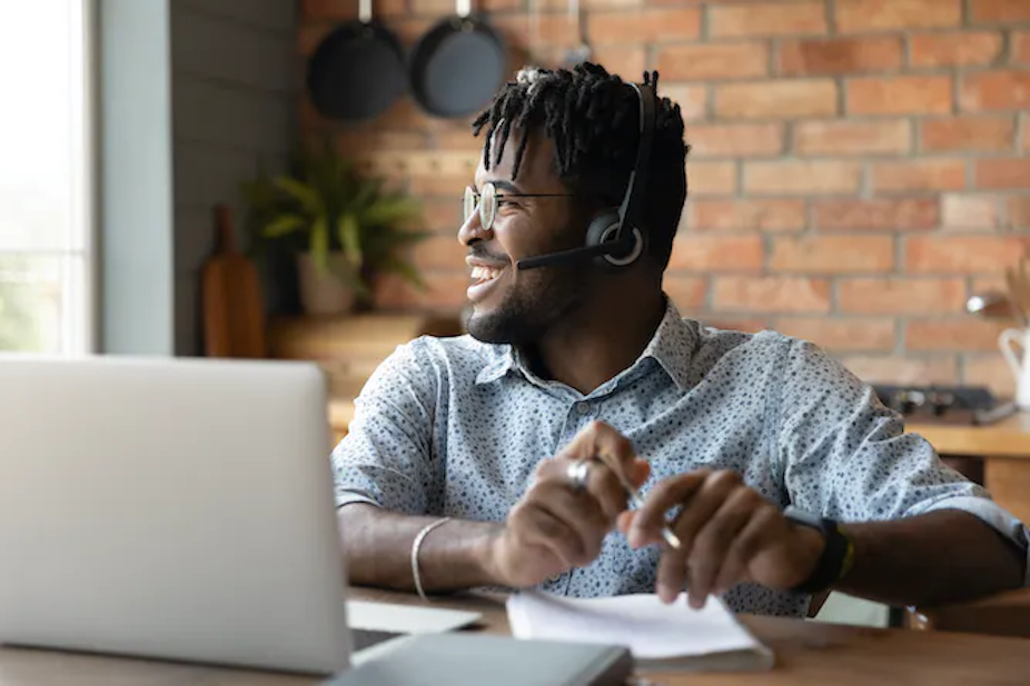 Seorang laki-laki yang memakai headphone duduk di meja dengan laptop, melihat ke samping, dan tersenyum.