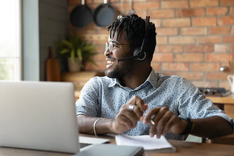 Seorang laki-laki yang memakai headphone duduk di meja dengan laptop, melihat ke samping, dan tersenyum.