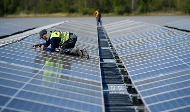 A man in a worker's vest crawls on his knees among a long line of solar panels floating on lake.