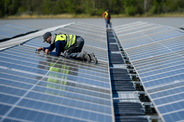 A man in a worker's vest crawls on his knees among a long line of solar panels floating on lake.