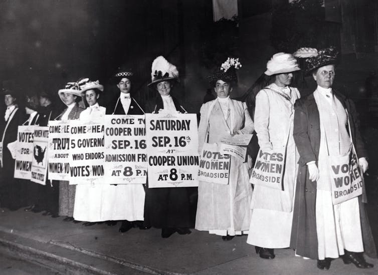 An historical photo of several women in fancy hats holding signs advertising a meeting about the right to vote.