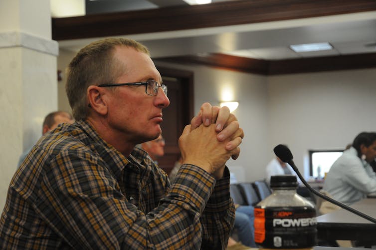 A man in a plaid shirt sits at a desk with a microphone, his hands folded as in prayer. He stares intently at whomever is speaking outside the image.