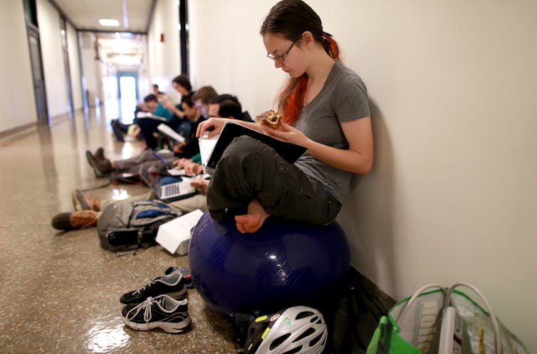 A female student sits on a yoga ball in a hallway reading a book. Other students are on laptops behind her.
