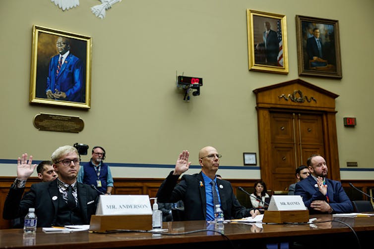 Three men dressed in suits and ties and seated at a long conference table raise their right hands as they are sworn in during a House Oversight Committee hearing. On the wall behind them hangs a portrait of the late U.S. Rep Elijah Cummings, chair of the House Oversight Committee.