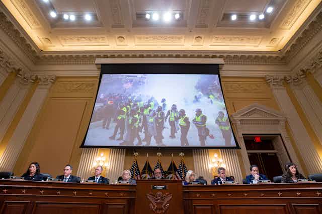 Nine people at a long desk in high-ceilinged meeting room, with a screen showing a photo of the Jan. 6 US Capitol riot.