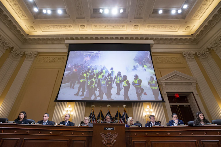 Nine people at a long desk in high-ceilinged meeting room, with a screen showing a photo of the Jan. 6 US Capitol riot.
