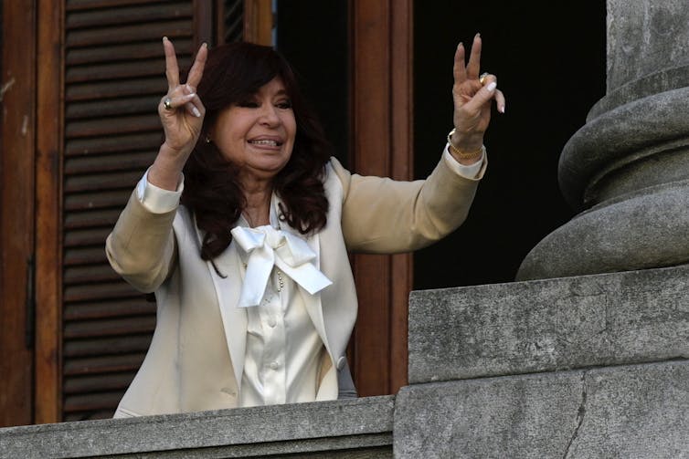 A woman wearing a beige jacket flashes a'v for victory' sign while standing on a balcony.