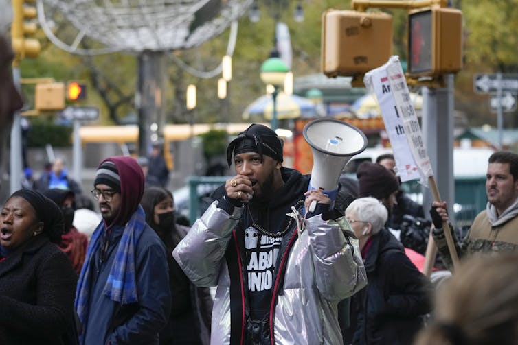 A man speaks into a megaphone in front of a crowd of protesters holding signs