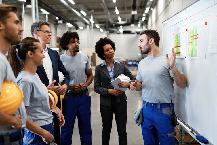 A group of people, some holding constriction hats, standing around a whiteboard having a discussion
