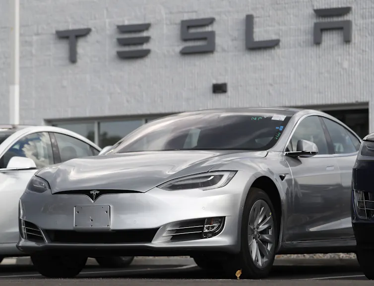 A silver car sitting outside a building with that says Tesla on the front