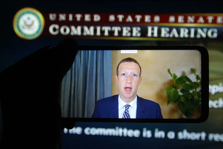 in the foreground a hand holds a smart phone displaying a video of a man in a business suit, and in the background a computer screen with a government logo across the top