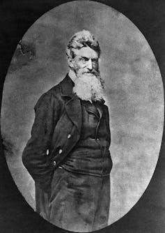 A black and white old-fashioned portrait of a standing man with a long white beard in black clothing.