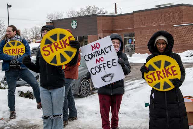Four people wearing coats hold up strike signs in the snow in front of a Starbucks