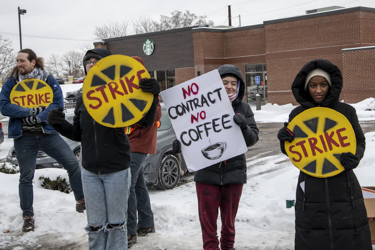Four people wearing coats hold up strike signs in the snow in front of a Starbucks