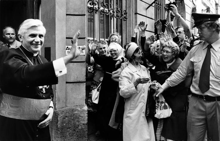 A young man dressed in priest robes waving to a group of parishioners.