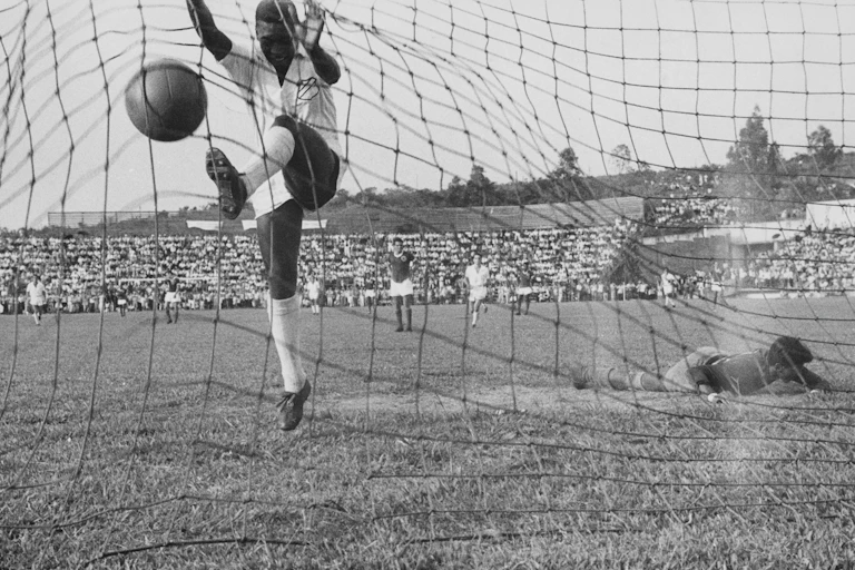 A soccer player in white kicks the ball into the net as the beaten goalkeeper lies on the ground.