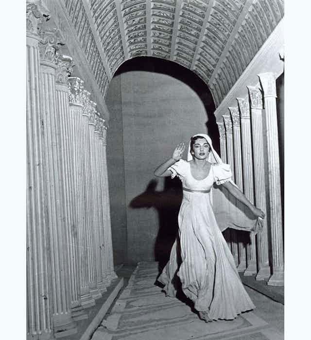 Una mujer con un vestido blanco se aproxima por un pasillo flanqueado de columnas.