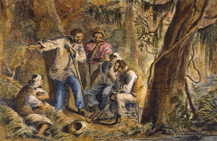 A small group of Black men and women listen to a Black man as they gather in the woods.