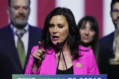 A dark-haired woman in a bright pink jacket speaks into a microphone.