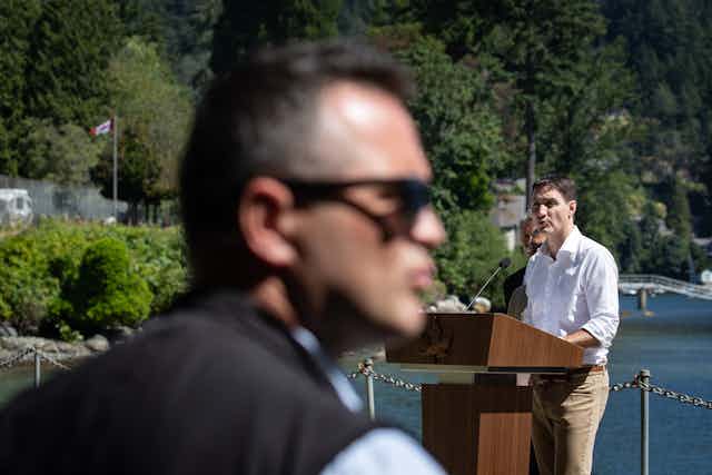 A man with sunglasses in the foregound as a man in a white shirt speaks into a microphone in the background. Trees and a Canadian flag are seen.