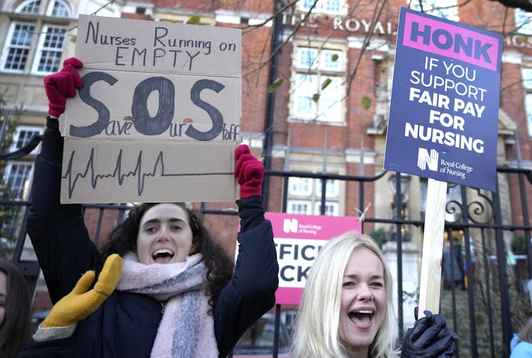 Dos mujeres blancas sostienen carteles, uno que dice "enfermeras corriendo sin carga, SOS", frente a un edificio de ladrillos