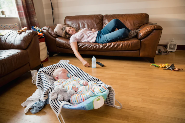 An exhausted mother sleeps on the sofa, baby in the foreground