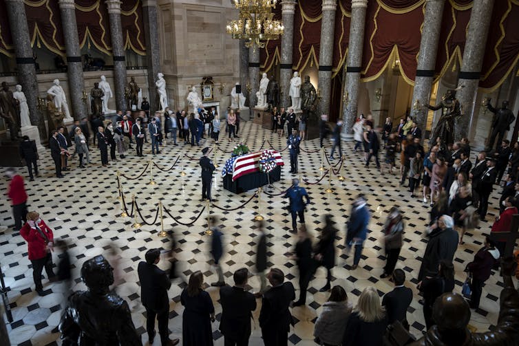 A flag-draped casket is in the middle of a large, stately hall, surrounded by people.