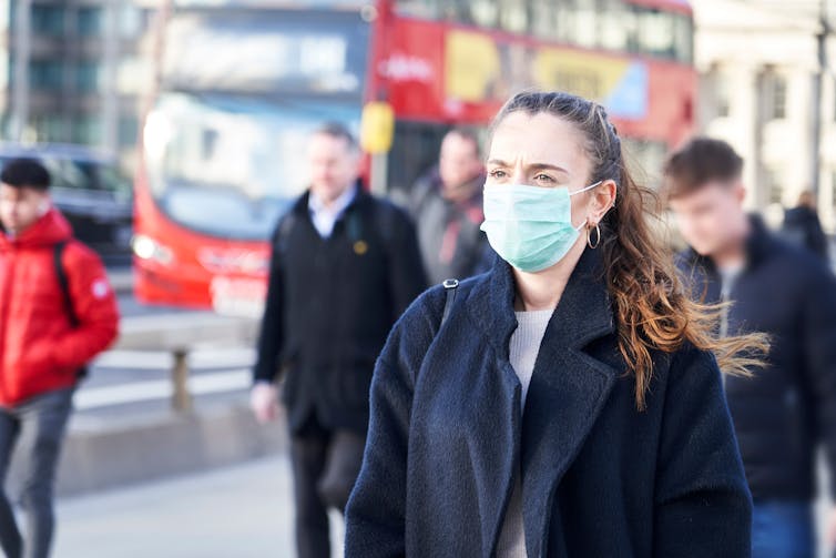A woman wearing a mask with people and a London bus in the background.