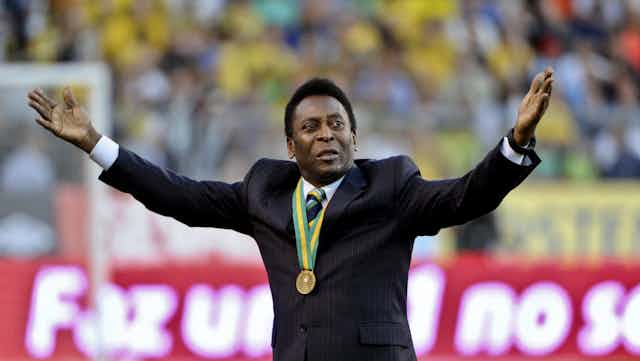 Pele stands in a stadium wearing a gold medal and holding both hands aloft. He's dressed in a black suit. 