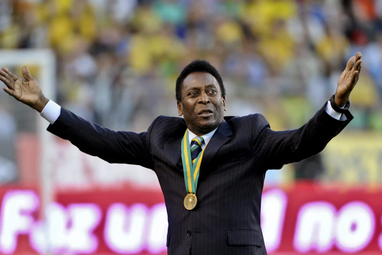 Pele stands in a stadium wearing a gold medal and holding both hands aloft. He's dressed in a black suit.
