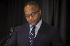 A Black man in a suit and glasses is seen at a news conference.