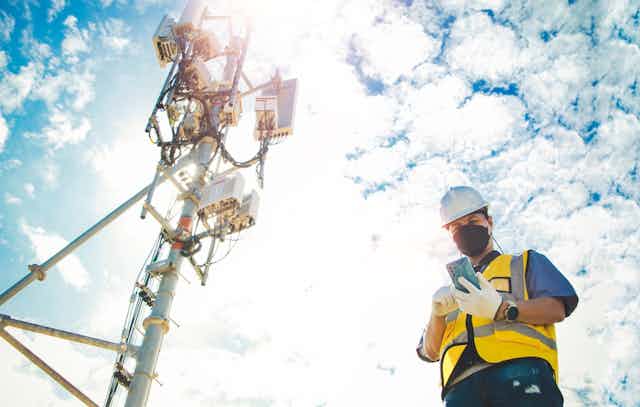 A man with face mask, helmet and high vis jacket checking his phone beside a 5G network antenna/telecommunications tower. Sun, cloud, blue sky background.