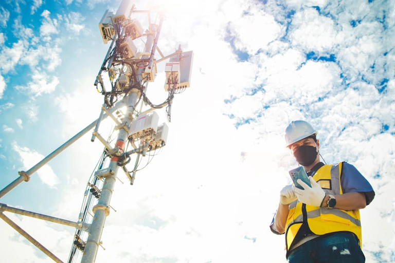 A man with face mask, helmet and high vis jacket checking his phone beside a 5G network antenna/telecommunications tower. Sun, cloud, blue sky background.