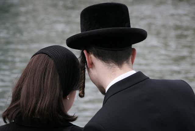 View from behind of an Orthodox Jewish man and woman, the woman is wearing a head covering and the man is wearing a black hat and payot