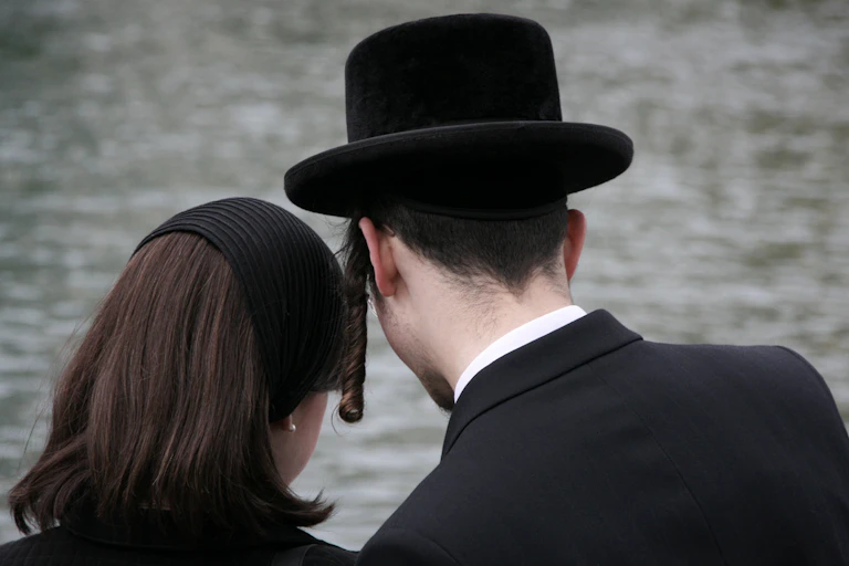 View from behind of an Orthodox Jewish man and woman, the woman is wearing a head covering and the man is wearing a black hat and payot