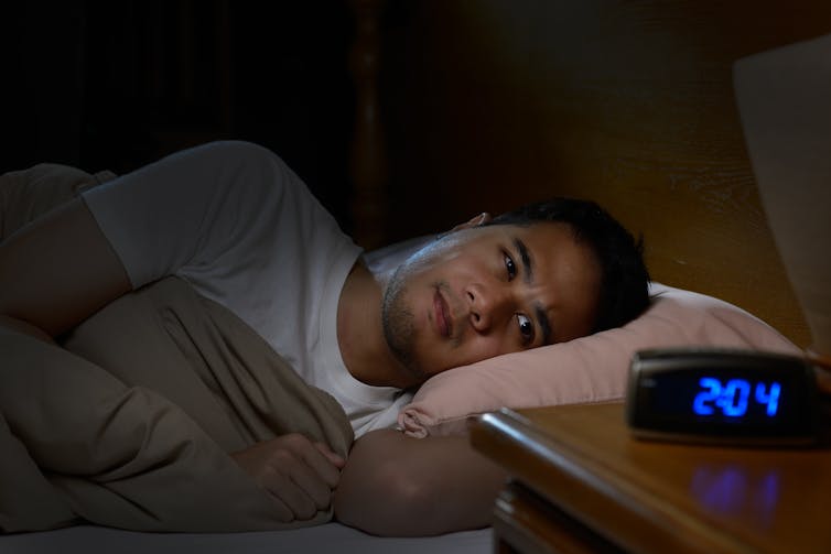 A man lies awake in bed. A clock on the bedside table shows 2:04.