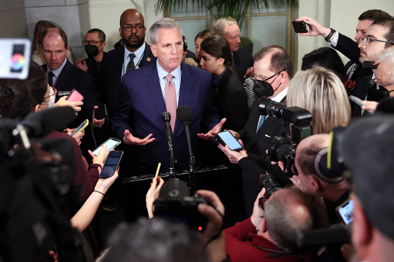 A man in a blue suit jacket and red tie is talking to lots of reporters holding out their phones to record him.