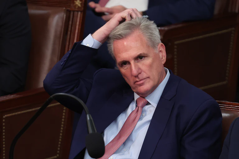 A gray-haired man in a blue blazer and red tie is sitting down and scratching his head.