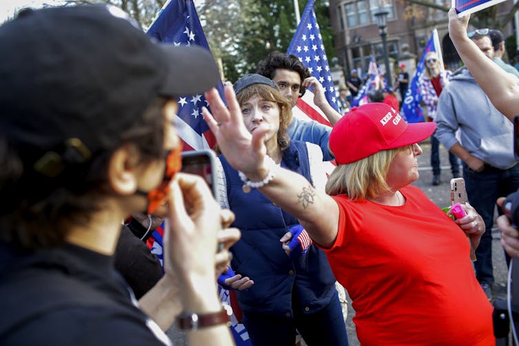 A middle-aged Caucasian woman in a red shirt and hat is reaching out to another Caucasian face with dark hair and a hat. Behind her stands a protester with an American flag.