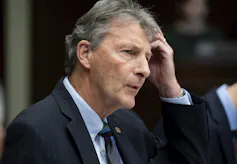 A man with thick grey hair has his hand to his forehead.