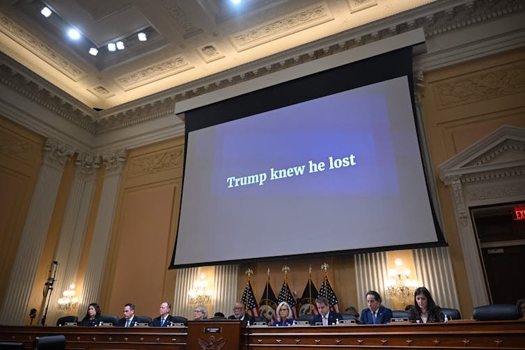 Representatives sit at a conference table. A screen overhead states: 'Trump knew he lost.'