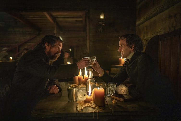 Christian Bale as Augustus Landor and Harry Melling as Edgar Allen Poe. The two men sit on opposite sides of a candlelit pub table and clink their glasses together in a cheers.