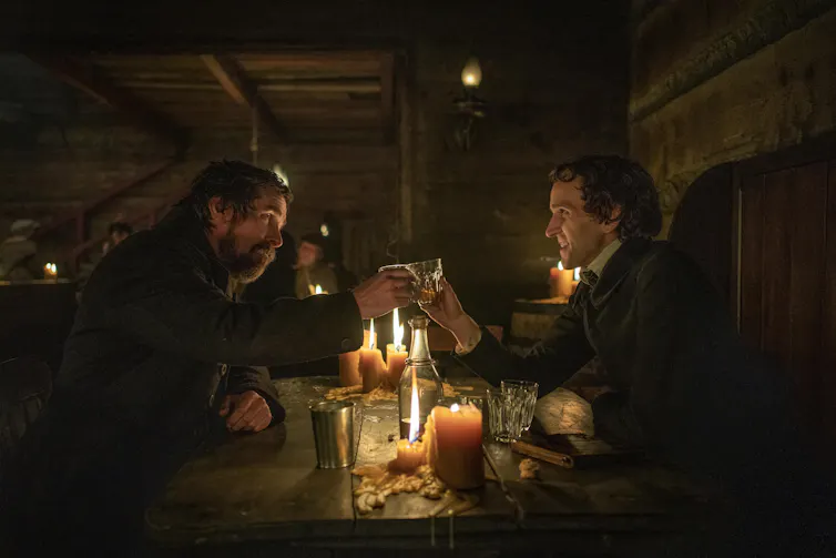 Christian Bale as Augustus Landor and Harry Melling as Edgar Allen Poe. The two men sit on opposite sides of a candlelit pub table and clink their glasses together in a cheers.